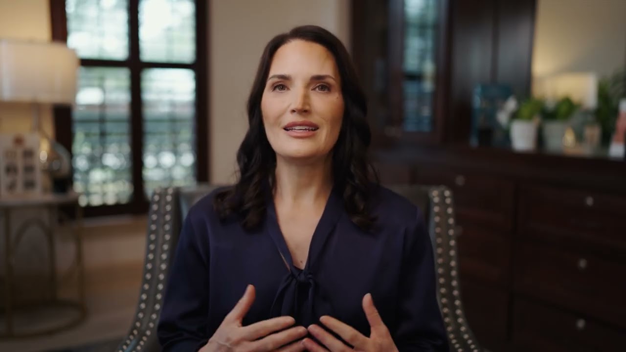Woman speaking indoors, wearing a dark blouse.