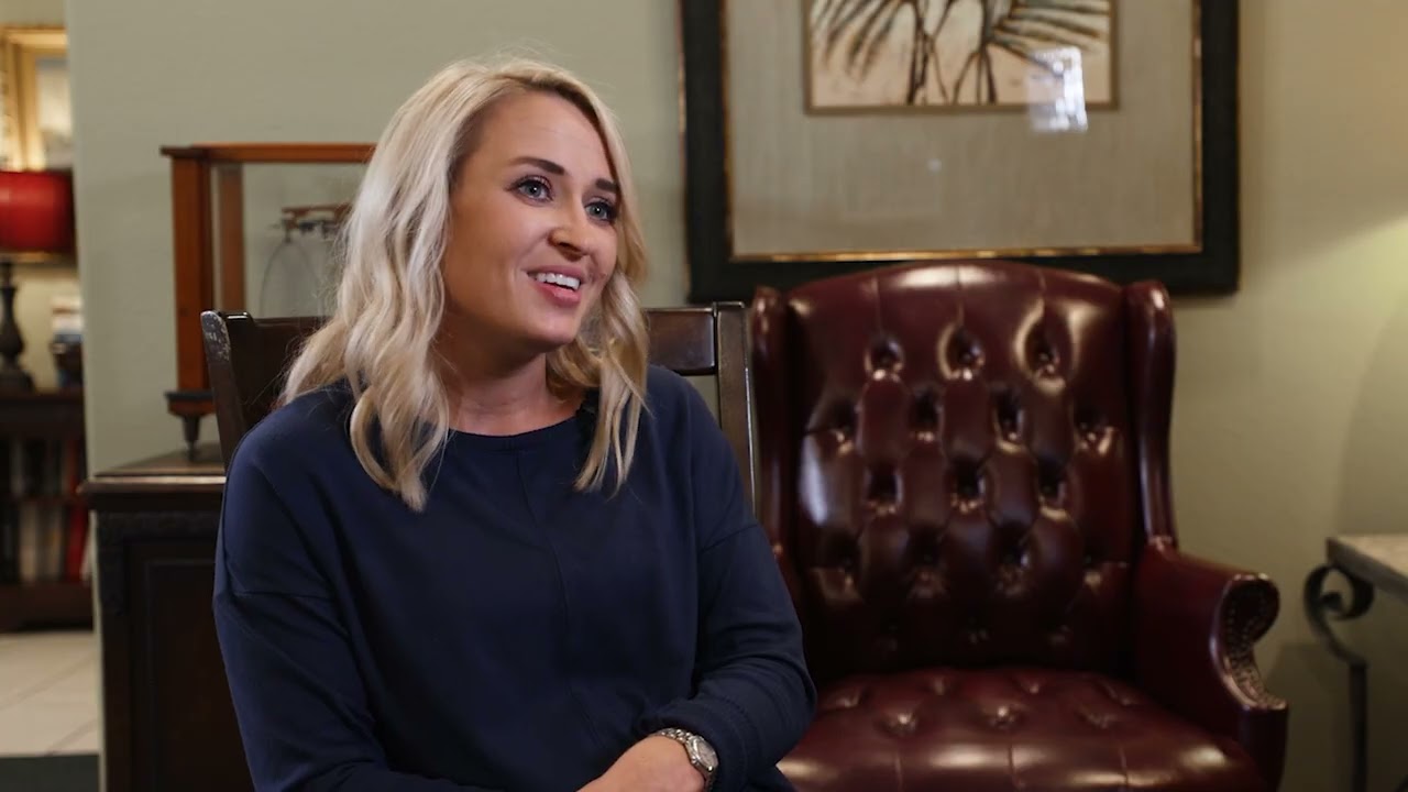 Dr. Abby Williams with a friendly smile is sitting in a cozy room on a dark leather armchair. She is wearing a dark blue top, and the background includes a framed artwork and warm-toned furnishings, creating an inviting atmosphere.